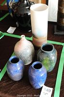 Five signed art pottery vases arranged on a dark cloth with green tape boundaries visible on the table. Includes blue vases, a tall ombre vase, a pinkish engraved vase, and a dark decorated vase.