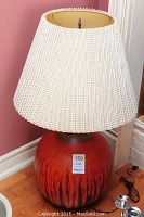 Full view of the mid century pottery table lamp showing the pottery base in reddish-orange with darker accents and a large woven cream shade.