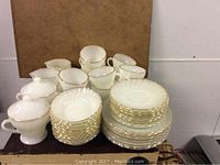 Front view showing stacks of white milk glass plates, bowls, cups, and covered sugar bowls with gold edging.