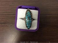 Front view of a handcrafted silver ring with an elongated turquoise stone in a purple display box showing the turquoise's matrix pattern and twisted rope setting.