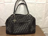 Front view of black and grey TNA tote bag with logo pattern, leather handles, and silver charm on front zip pocket.