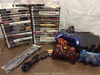 Photo showing the black PlayStation 2 slim console, three wired controllers (blue, translucent red, translucent blue), AV and power cables, and 41 PS2 game cases.