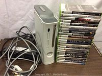 Xbox 360 white console standing upright next to a stacked set of 24 Xbox 360 game cases, power supply and AV cables in view.