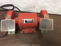 Front-facing photo showing the red Ashland double bench grinder with central motor housing, 5-inch grinding stones, and attached clear protective eye shields.