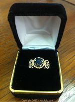 Ring displayed inside a black velvet jewelry box showing full ring from the front with visible large central blue stone and gold-tone metal band.
