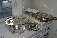 Overview showing ornate covered entrée dish on right, oval tray on left, two round bowls front center, and silver rack with ceramic insert on top right.