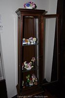 Full view of closed wooden curio cabinet with glass shelves containing several miniature fine bone china floral figurines.