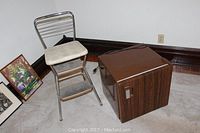 Small wooden mini refrigerator and Cosco metal step stool shown together in corner of room with framed artwork nearby.