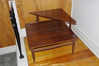 Full view of the Mersman mid century side table showing two tiers, triangular top tier and square lower tier, with tapered legs on hardwood flooring against wood paneled wall.