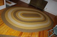 Full view of an oval braided area rug in tan, brown, beige, black, and mustard colors on wooden floor near radiator.