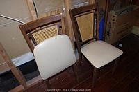 Two wood frame folding chairs with caned backs and cream vinyl upholstered seats placed side by side