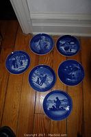 Six blue-and-white Royal Copenhagen collector plates laid out on the floor, showing detailed winter and Christmas scenes.