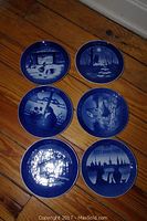 Six Royal Copenhagen collector plates arranged on wooden floor showing various winter and holiday themed blue and white scenes.