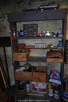 Shelf with multiple boxes containing various bolts, nuts, screws, cans of liquid chemicals and paint, cardboard boxes with miscellaneous hardware and tools.