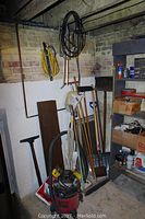 Photo showing household tools including hoe, shovel with wooden handles, broom, vacuum cleaners, and laminate shelves against basement wall.