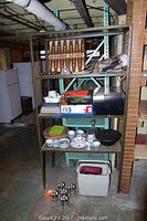 Photo of multi-tier metal shelving unit with game boxes on top, decor gun, various china pieces including cups, saucers, and bowls, a silverplate basket, black and red food warmer in original box, cooler, and metal drink holders on the floor.
