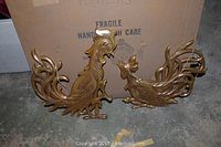 Two brass or bronze rooster wall plaques with detailed feather design, laid against boxed background on floor.