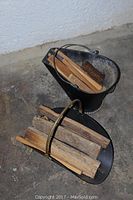 Photo showing black metal coal bucket and black metal wood basket both filled with pieces of firewood on concrete floor.
