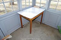 Full view of rectangular wood frame table with white enamel top placed in a sunroom. Enamel top shows some visible marks and wear.