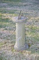 Concrete sundial on pedestal standing on grass, showing full height and textured concrete base.