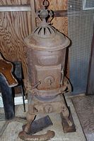 Full view of rusty heavy globe oak pot belly stove with ornate vented top, front door, and spring handle. Visible damage with broken side piece.