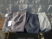 Three pairs of men's dress slacks in varying shades of gray displayed on table, showing pleated fronts and sizes on tags.