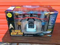 Front view of the sealed boxed set of 7 Classic Star Trek TOS bridge crew action figures including Kirk, Spock, McCoy, Chekov, Sulu, Uhura, and Scotty with captain's chair on bridge background.