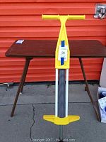 Full view of the yellow Sport Fun Inc. pogo stick with yellow plastic handle and footrests, metal spring, and standing upright outdoors in front of a red background.