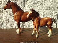 Side view of the brown Byer Clydesdale mare standing beside the foal on a wooden surface with a white textured wall background.