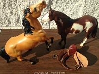 Two model horses on a wooden surface against a white textured background, showing the rearing yellow Mustang with black mane and the brown and white paint horse alongside a small leather saddle.