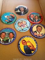 Seven political inauguration pins laid out on a tan surface including Bill Clinton, Hillary Clinton, Al Gore, and John F. Kennedy. Pins show some scuffs and wear.