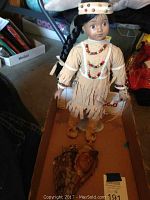 Full view of Native American style doll showing leather dress with fringe, bead necklace, headdress, and sandals. Doll standing on cardboard box.
