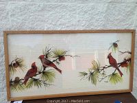 Front view of original glass painting showing a horizontal composition of red cardinals on pine branches, in light wood frame against a white textured wall.