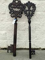 Two large decorative metal wall hanging keys shown upright against an exterior white wall. Both keys have detailed ornate bows and decorative bits, with a dark brown distressed finish.