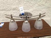 Top view of the wall light fixture showing three frosted bell-shaped glass shades and curved brushed metal frame, placed on a table against a wall.