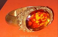 Close-up angled view of the bangle showing the large amber-colored faceted stone and ornate swirl detailing on the gold-tone metal setting and band.