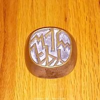 Top view of a small rounded rectangular solid brass paperweight featuring a silver-toned embossed geometric abstract design on top, placed on a wood surface.