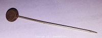 Stick pin with small Indian Head coin mounted at end, showing profile and feather headdress, set on a flat surface.