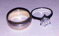 Photo of two sterling silver rings side by side on a plain background, showing both the ladies ring with a large round cubic zirconia and the men's solid band.