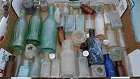 Top-down photo of an assortment of old glass bottles in a box showing variety of green, clear, brown, and blue bottles, some with corks and embossed markings.