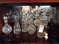 Multiple cut crystal decanters and stemmed glasses arranged on wooden shelf.