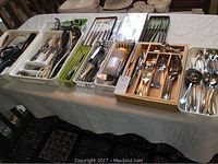 Tabletop display of multiple trays and containers filled with assorted cutlery and kitchen tools, showing the large quantity and variety of items.