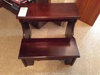 Front view of dark wooden two-step stool with curved sides and solid steps on carpeted floor.