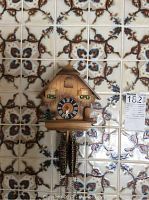 Front view of wooden German cuckoo clock with two pine cone weights hanging beneath dial, against decorative tile background