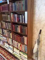 Photo shows a tall wooden bookcase filled with many books of various sizes and colors against a wallpapered wall.