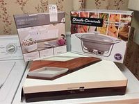 Three kitchen serving items still in packaging: 7-piece white porcelain serving set, white casserole warmer with metal frame, wooden appetizer tray box.