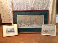 Three framed art pieces grouped on floor: snowy landscape, panoramic townscape, coastal boats