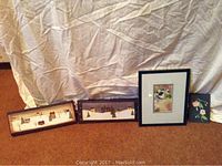 Four wall art pieces arranged on carpeted floor against fabric backdrop: two long rectangular painted village scenes, one floral print and one framed colorful print.