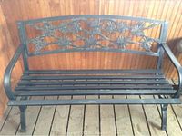 Front and side view of black metal garden bench with floral pattern on the backrest and slatted seat on wooden deck floor.