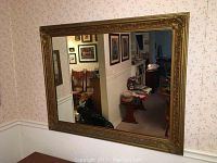 Full view of the large rectangular vintage wall mirror hanging on a floral patterned wall, showing the entire carved gold wood frame and reflective glass surface.